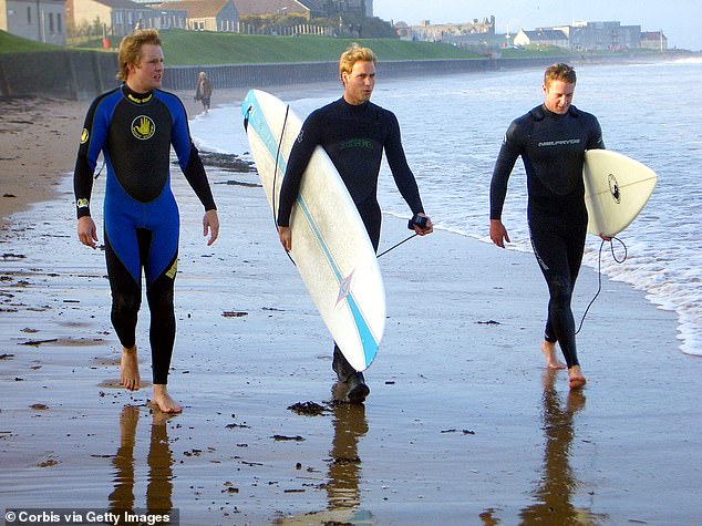 Prince William in wetsuit carrying a surfboard along the shoreline with two friends at St Andrews in Scotland, 2004